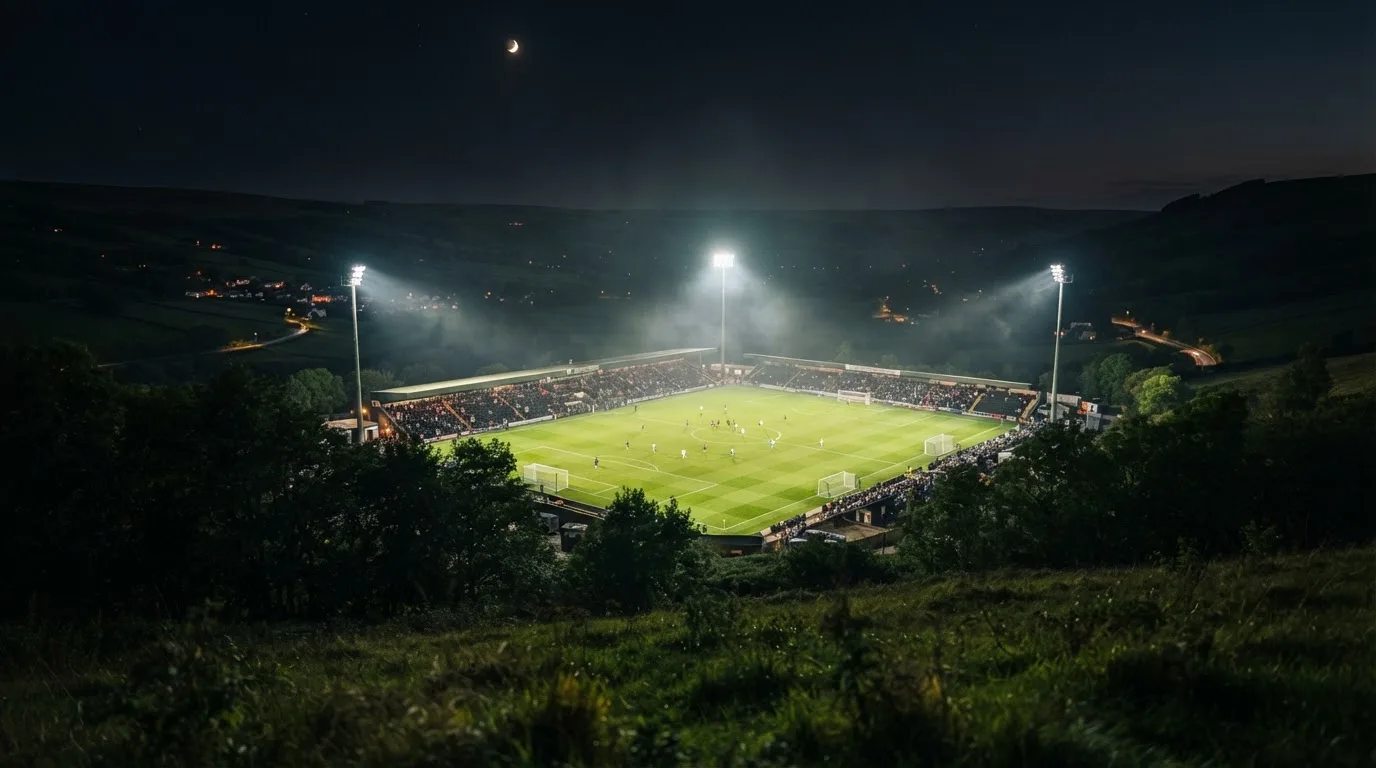 Ein Stadion unter Flutlicht bei Nacht, von einem Hügel aus der Ferne betrachtet, mit Dunst über dem Spielfeld.