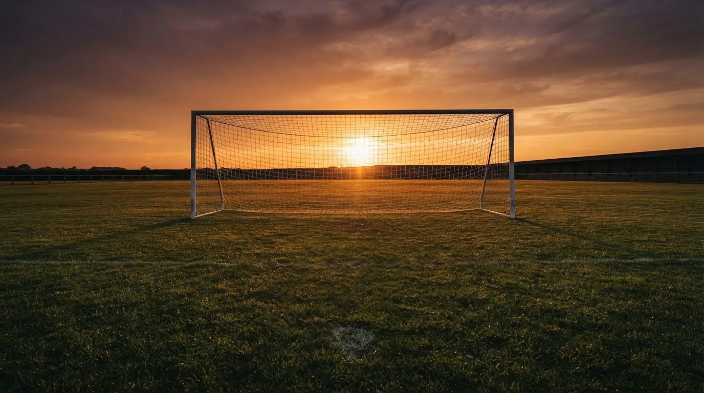 Ein Fußballtor mit Netz bei Sonnenuntergang auf einem leeren Platz, die Schatten der Torpfosten liegen lang auf dem Gras.