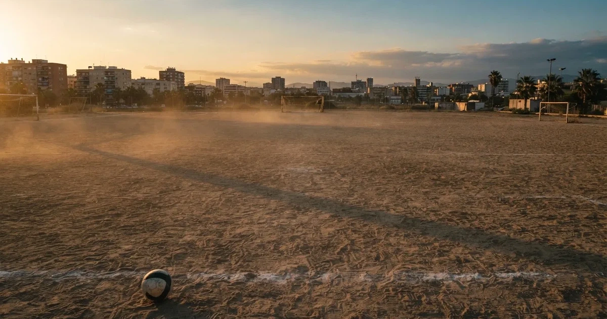Ein sandiger Fußballplatz am Rand einer Stadt in warmem Abendlicht, Staub in der Luft und zwei Tore in der Ferne.