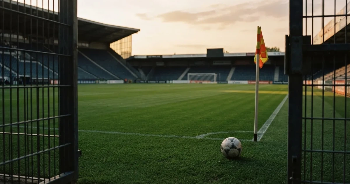 Blick durch ein Stadiontor auf einen grünen Rasen im Abendlicht, ein Ball liegt an der Eckfahne, die Ränge sind leer.