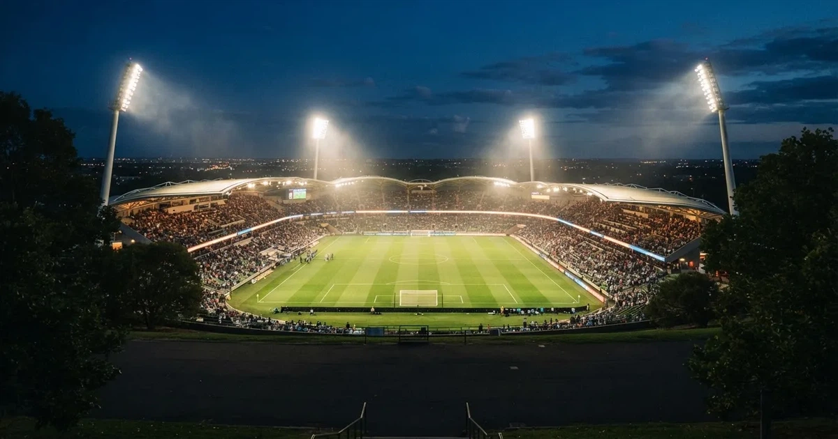 Ein großes Stadion bei Nacht, aus der Ferne unter Flutlicht gesehen, die Ränge hell erleuchtet, der Rasen leuchtend grün.