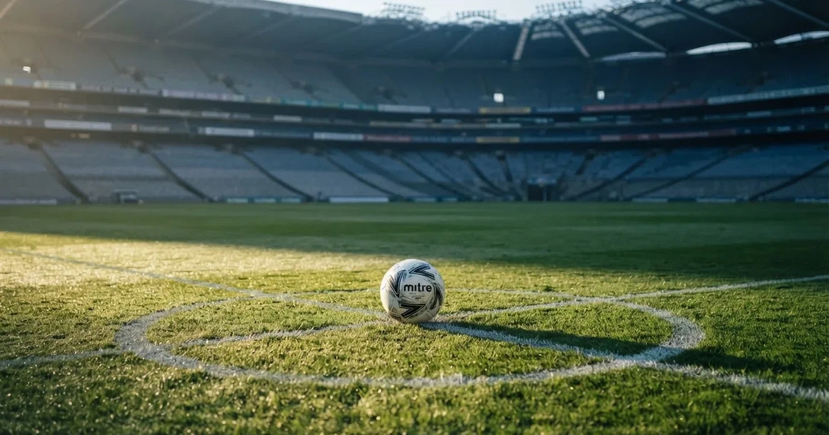 Ein Fußball liegt auf dem Mittelkreis eines Stadionrasens am frühen Morgen, im Hintergrund leere Ränge ohne erkennbare Beschriftung.