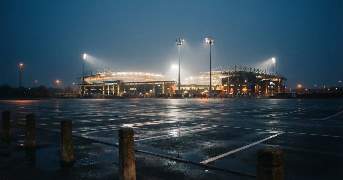Ein Stadion unter Flutlicht aus großer Entfernung gesehen, davor ein regennasser Parkplatz in der Dämmerung.