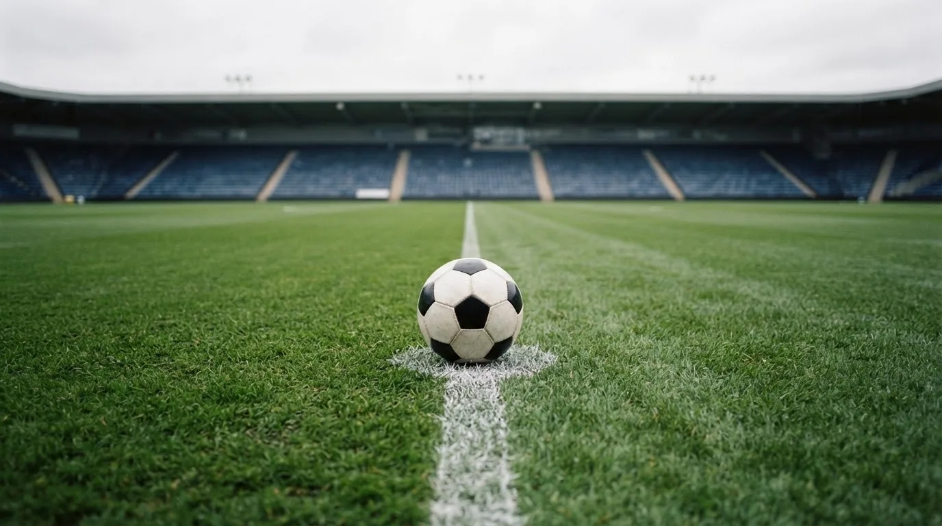 Ein Fußball liegt auf dem Anstoßpunkt eines leeren Rasenplatzes, im Hintergrund eine ruhige Tribüne unter bedecktem Himmel.