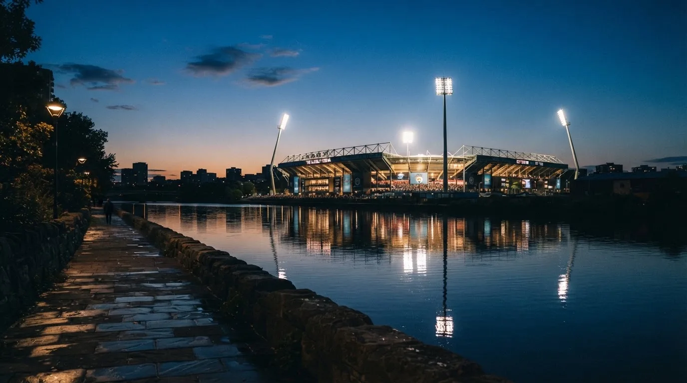 Ein Stadion unter Flutlicht aus der Ferne gesehen, im Vordergrund ein ruhiger Fluss und ein Fußweg.