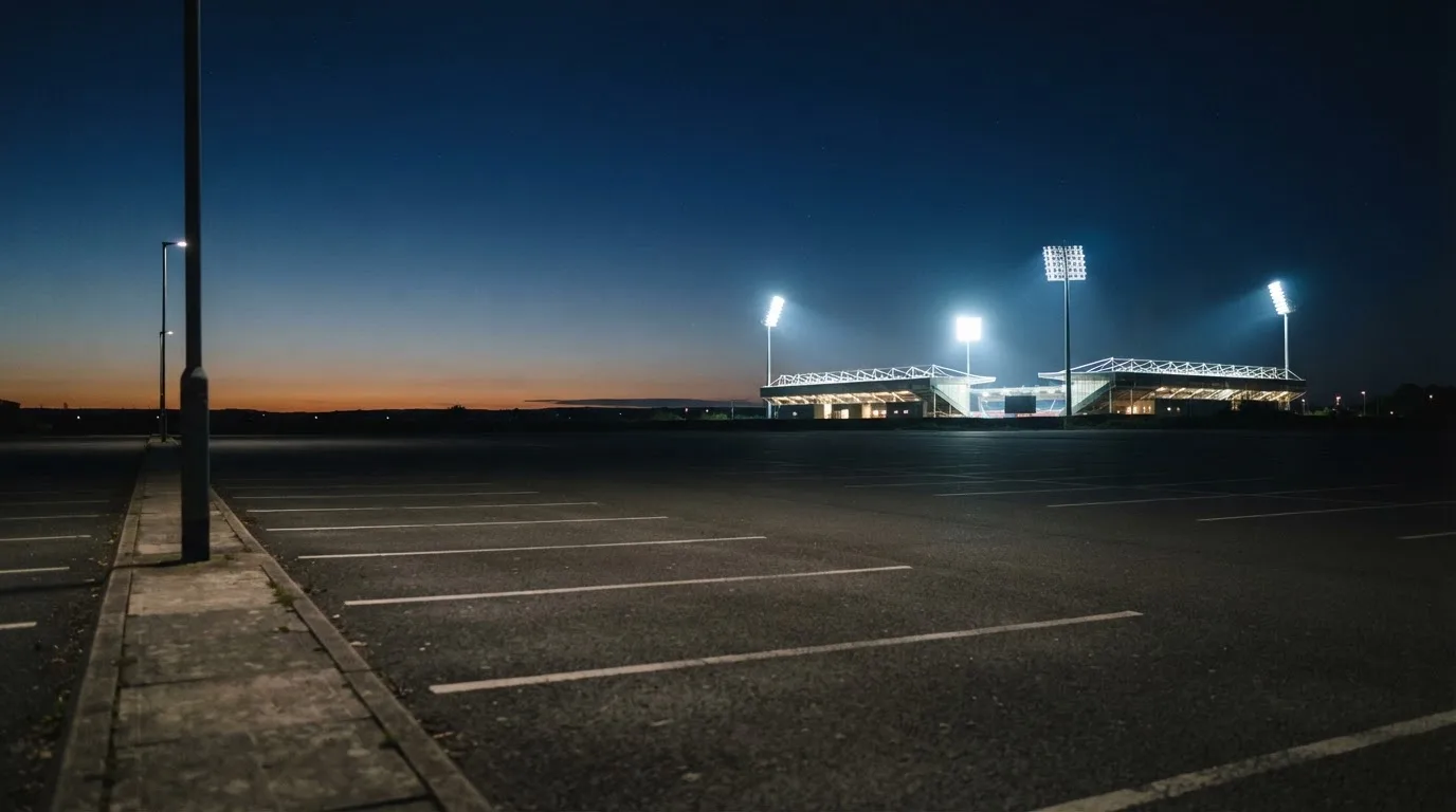 Ein Stadion unter Flutlicht aus der Ferne gesehen, davor ein leerer Parkplatz in der Dämmerung.