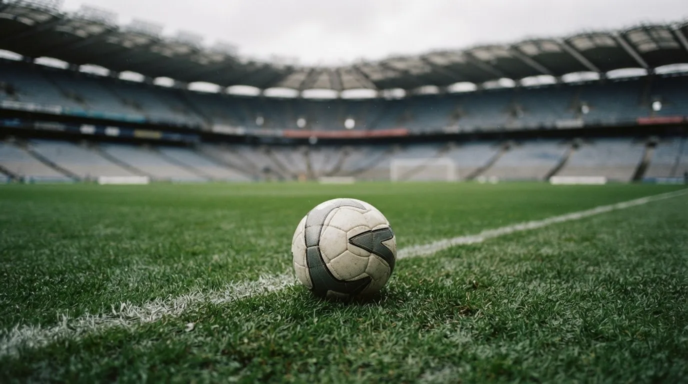 Ein Fußball auf einem Rasen, im Hintergrund ein leeres Stadion bei bewölktem Himmel.