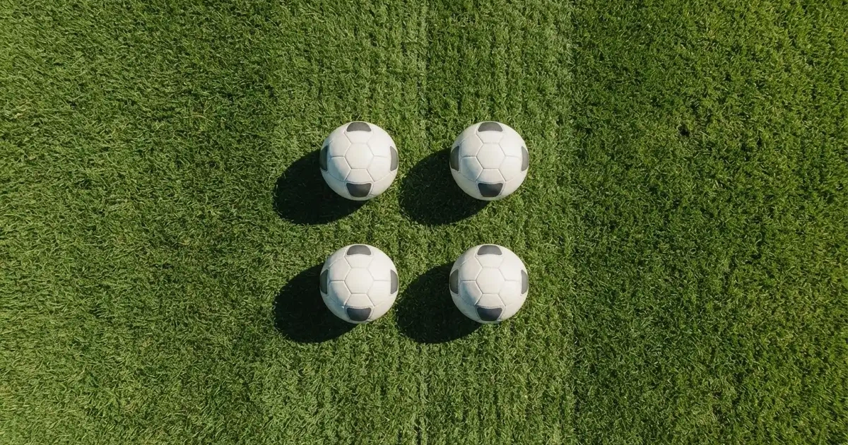 Vier Fußbälle in einem Quadrat auf einem Rasenplatz, von oben fotografiert, mit harten Mittagsschatten.