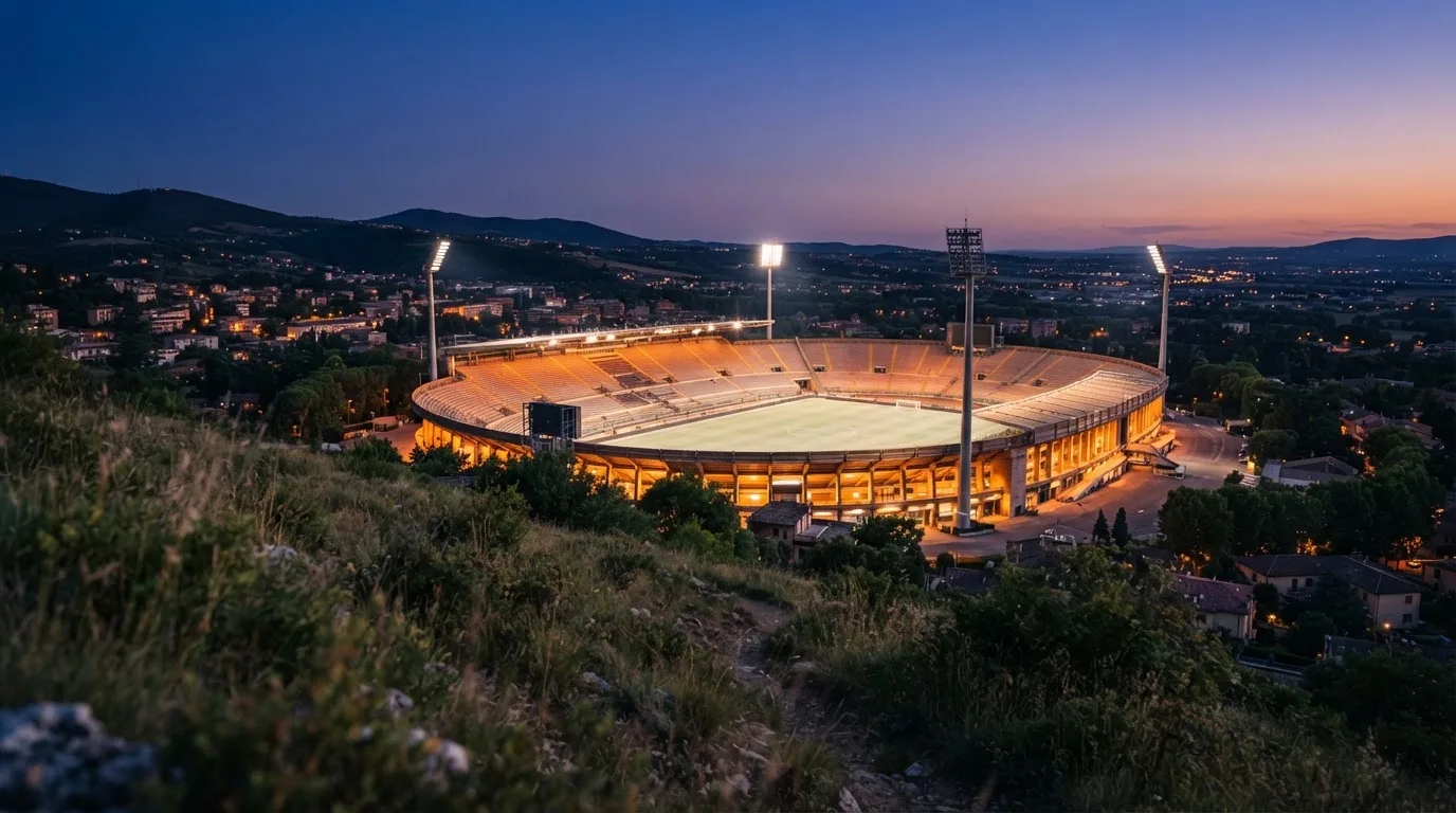 Ein leeres Stadion mit hell erleuchteten Rängen in der Abenddämmerung, von einem Hügel aus fotografiert.