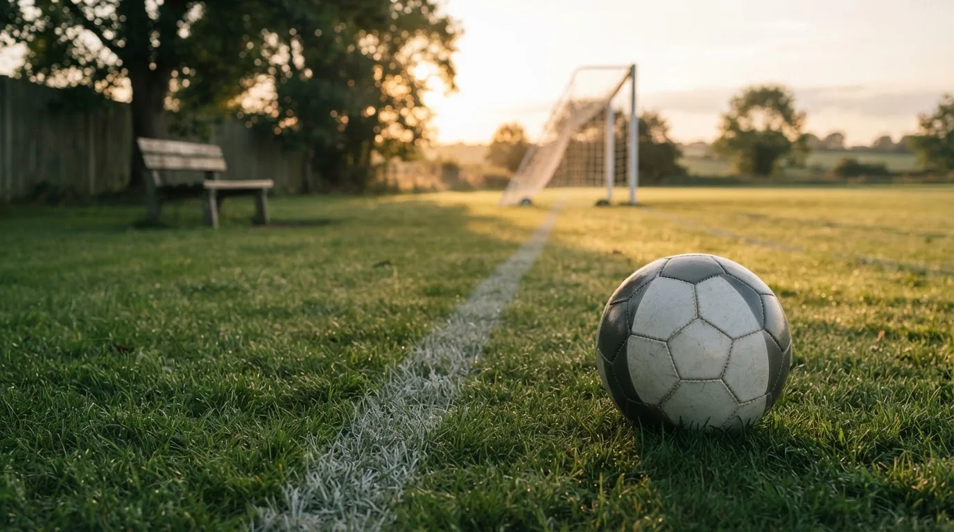 Ein Fußball liegt im Gras am Spielfeldrand, dahinter eine leere Bank und ein Tor im Gegenlicht.