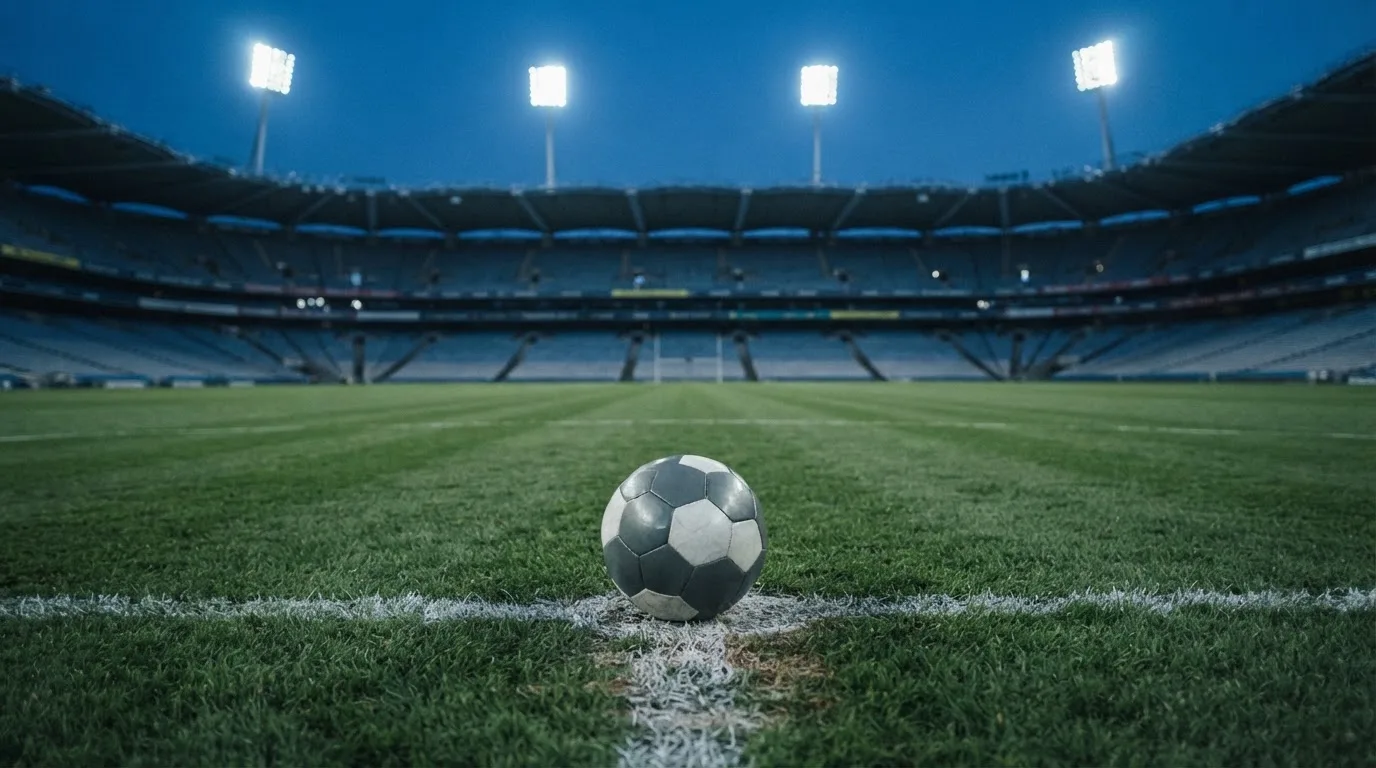 Ein Fußball liegt auf dem Anstoßpunkt eines leeren Rasenplatzes, im Hintergrund ein Stadion unter Flutlicht in der Abenddämmerung.