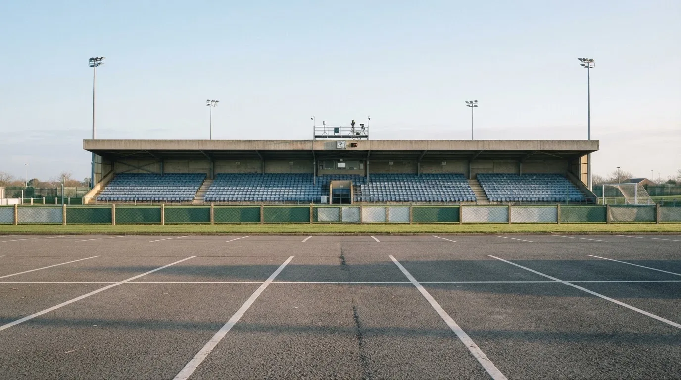Eine leere Tribüne eines kleinen Stadions am frühen Morgen, aus der Ferne über einen menschenleeren Parkplatz fotografiert.