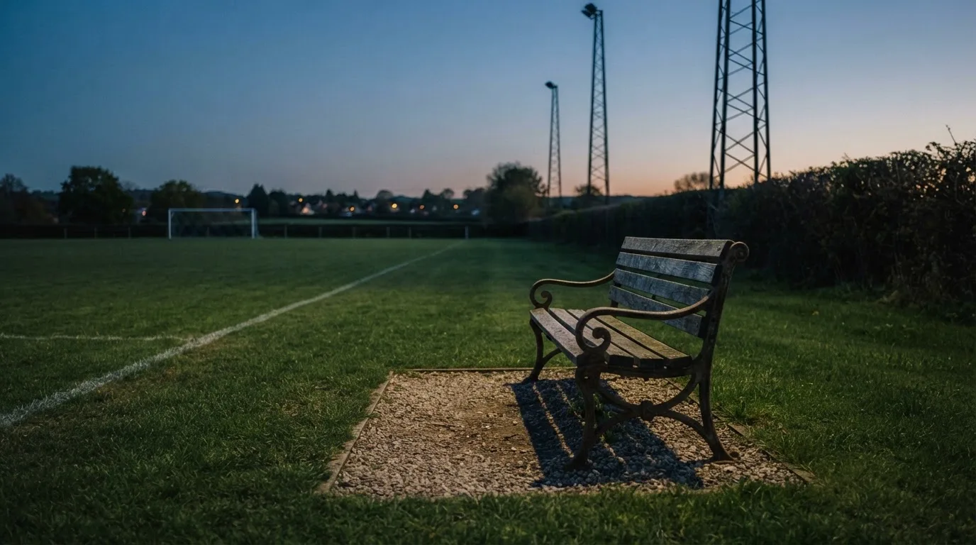 Eine leere Parkbank am Rand eines Fußballfelds in der Abenddämmerung, im Hintergrund ragen dunkle Flutlichtmasten in den Himmel.