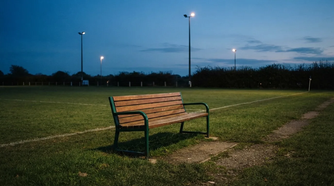Eine leere Parkbank am Rand eines Fußballfelds in der Abenddämmerung, im Hintergrund ragen dunkle Flutlichtmasten in den Himmel.