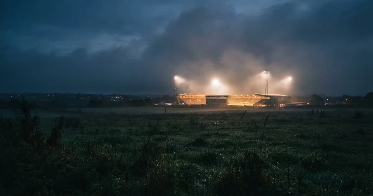 Ein Stadion unter Flutlicht, aus großer Entfernung über eine dunkle Wiese fotografiert, am Himmel zieht Nebel auf.
