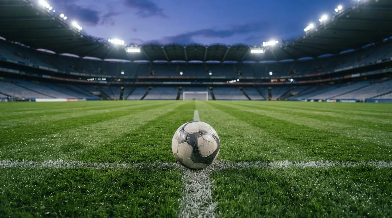 Ein Fußball liegt auf dem Anstoßpunkt eines leeren Rasenplatzes, im Hintergrund steht ein Stadion unter Flutlicht in der Dämmerung.