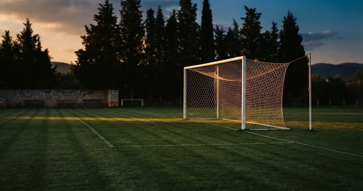 Ein Fußballtor mit Netz auf einem leeren Platz im Abendlicht, der Rasen frisch gemäht, im Hintergrund Zypressen.