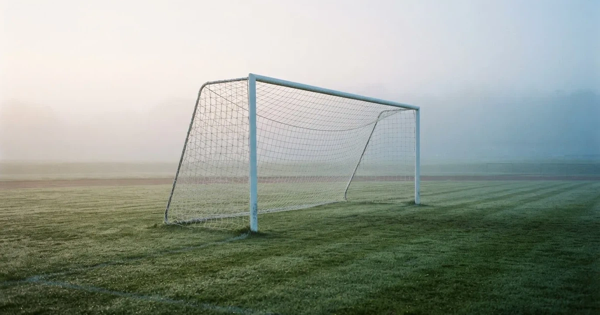 Ein Fußballtor mit Netz steht auf einem leeren Platz im Nebel der Morgendämmerung, der Rasen ist taufeucht.