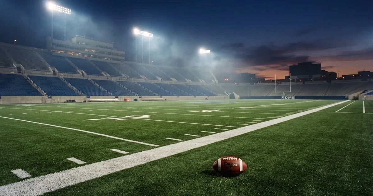 Ein Footballstadion bei Flutlicht aus der Ferne gesehen, davor ein leerer Rasen mit Yard-Linien und ein ovaler Ball am Spielfeldrand.