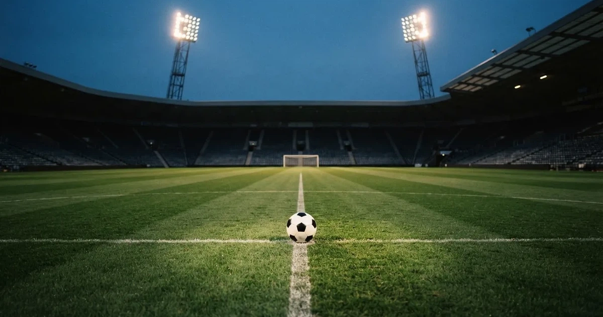 Ein Rasenplatz in der Dämmerung mit eingeschalteten Flutlichtmasten, ein Ball liegt am Anstoßpunkt, die Tribünen im Hintergrund sind unscharf.