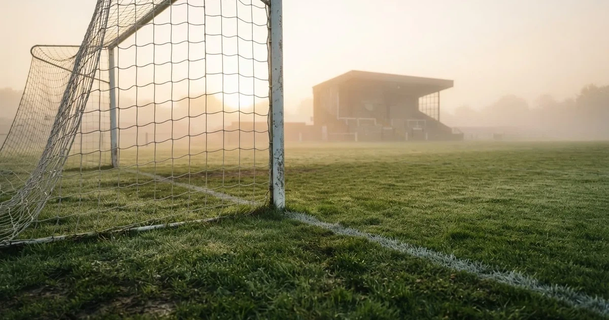 Ein Fußballtor mit Netz steht im Morgennebel auf einem leeren Rasenplatz, im Hintergrund die Silhouette einer kleinen Tribüne.