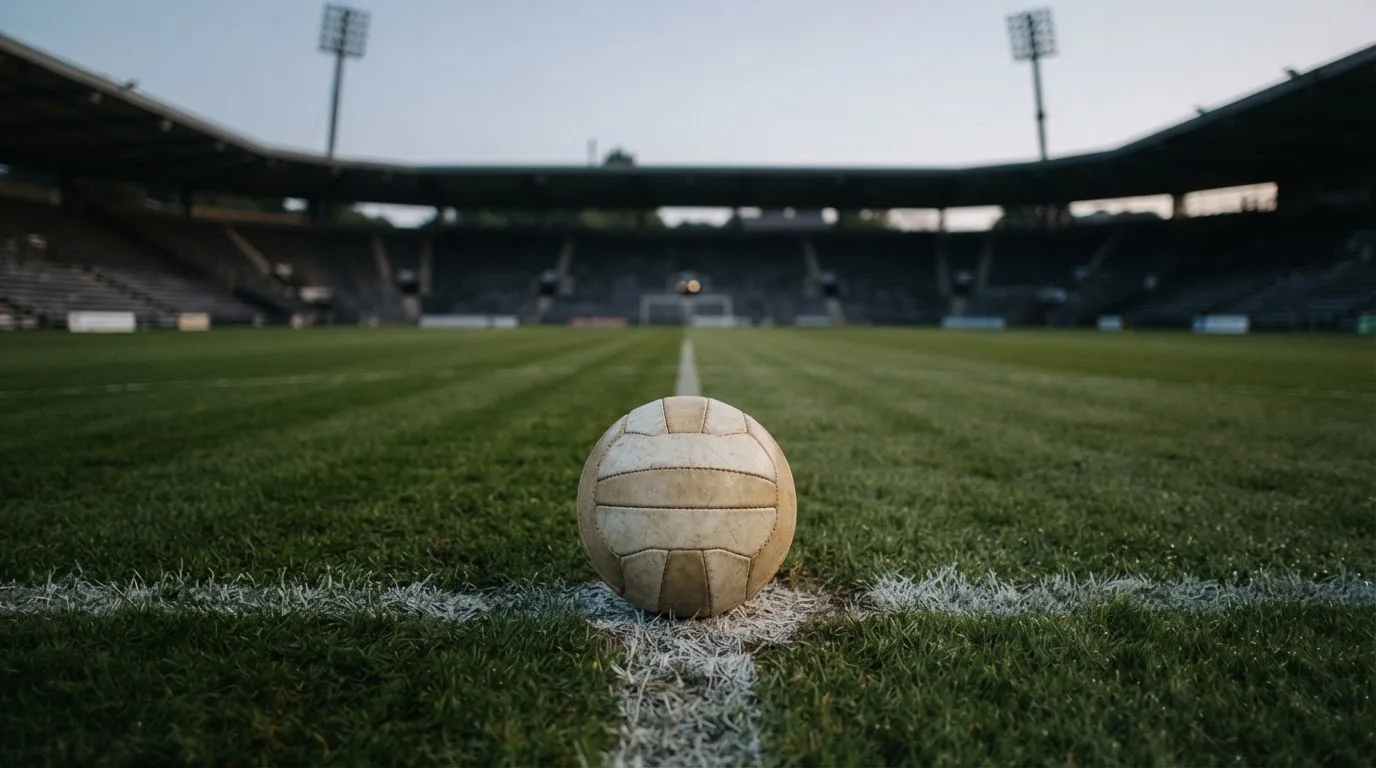 Ein Fußball liegt auf dem Anstoßpunkt eines leeren Rasenplatzes, dahinter ragen dunkle Ränge in den Abendhimmel.