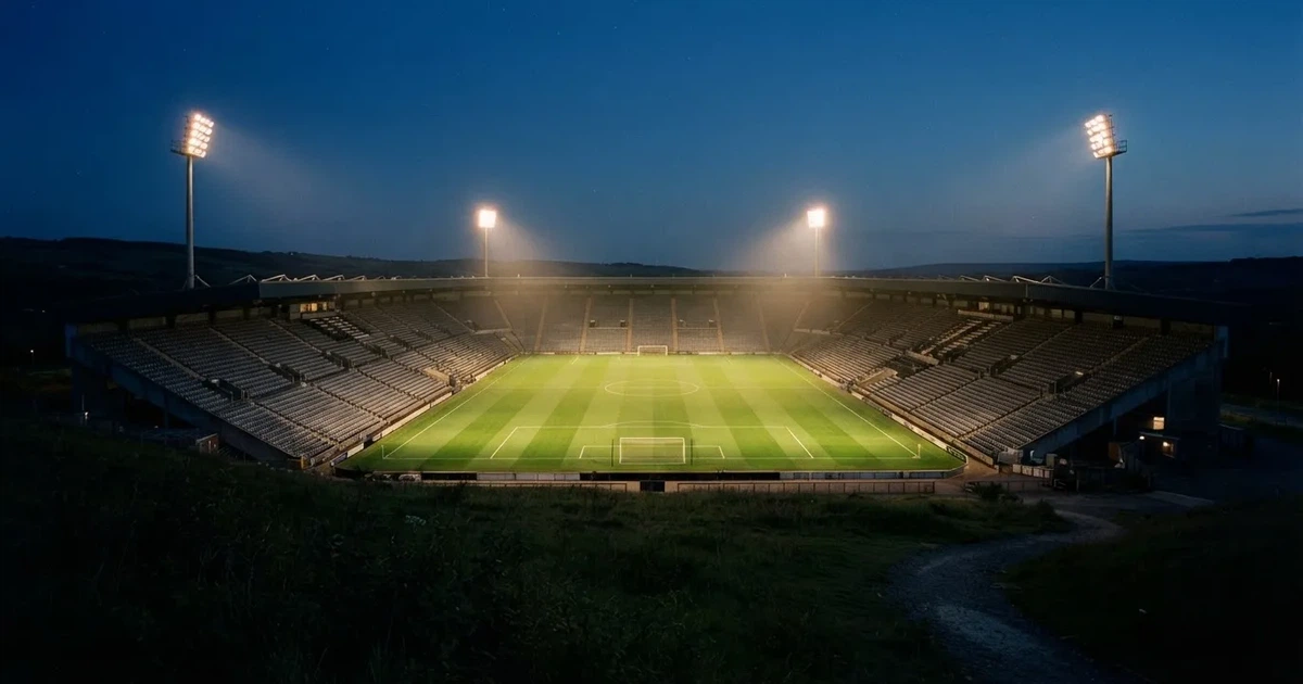 Ein Fußballstadion am Abend unter Flutlicht aus der Ferne gesehen, die Tribünen sind leicht beleuchtet und der Himmel dunkelblau.
