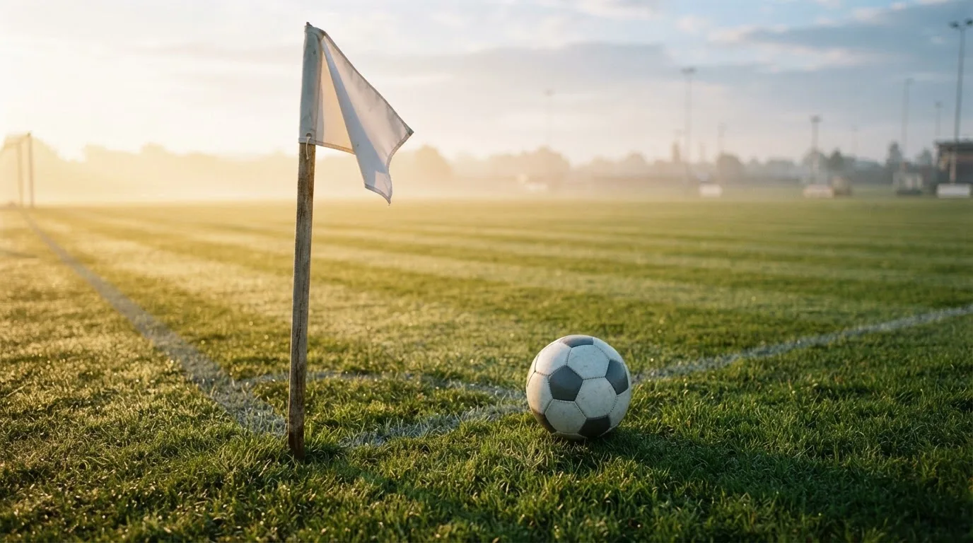 Ein Fußball liegt auf Rasen neben einer Eckfahne ohne Aufdruck auf einem leeren Platz im Morgenlicht.