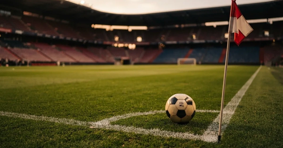 Ein Fußball liegt am Viertelkreis an der Eckfahne eines Rasenplatzes, im Hintergrund leere Tribünen in warmem Abendlicht.
