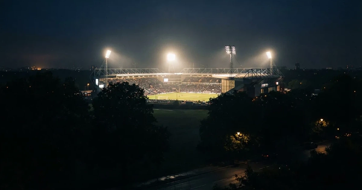 Ein Fußballstadion bei Nacht aus großer Entfernung, hell erleuchtet unter Flutlicht, davor eine dunkle Straße und Bäume.