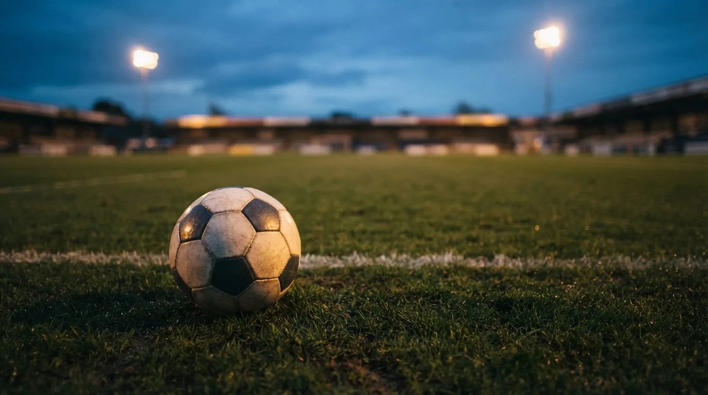 Ein Fußball liegt auf einem Rasenplatz in der Dämmerung, im Hintergrund ein ferner Stadionrand unter Flutlicht.